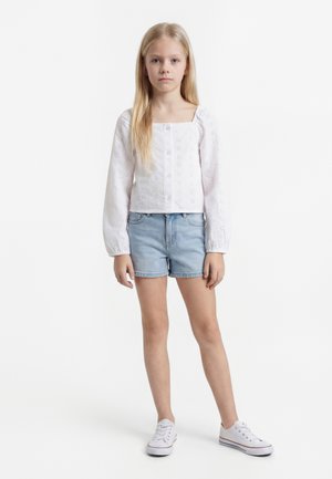Ung jente med langt blondt hår iført en hvit knappet bluse med hullmønsterdetaljer, lyse denimshorts og hvite sneakers, står.