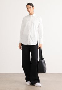 Camicia bianca con bottoni e decorazioni testurizzate, abbinata a pantaloni neri a gamba larga. Una borsa nera e scarpe bianche completano l'outfit.