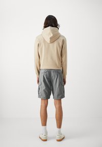 Sudadera beige con capucha y bolsillo canguro, combinada con pantalones cortos de cargo grises con bolsillos laterales. Calcetines blancos y zapatillas con logotipo.