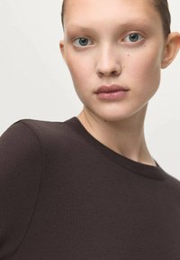 Hnedý pletený top s okrúhlym výstrihom. Tkanina sa zdá byť hladká a tesne priliehajúca, zdôrazňujúca obrysy odevu.