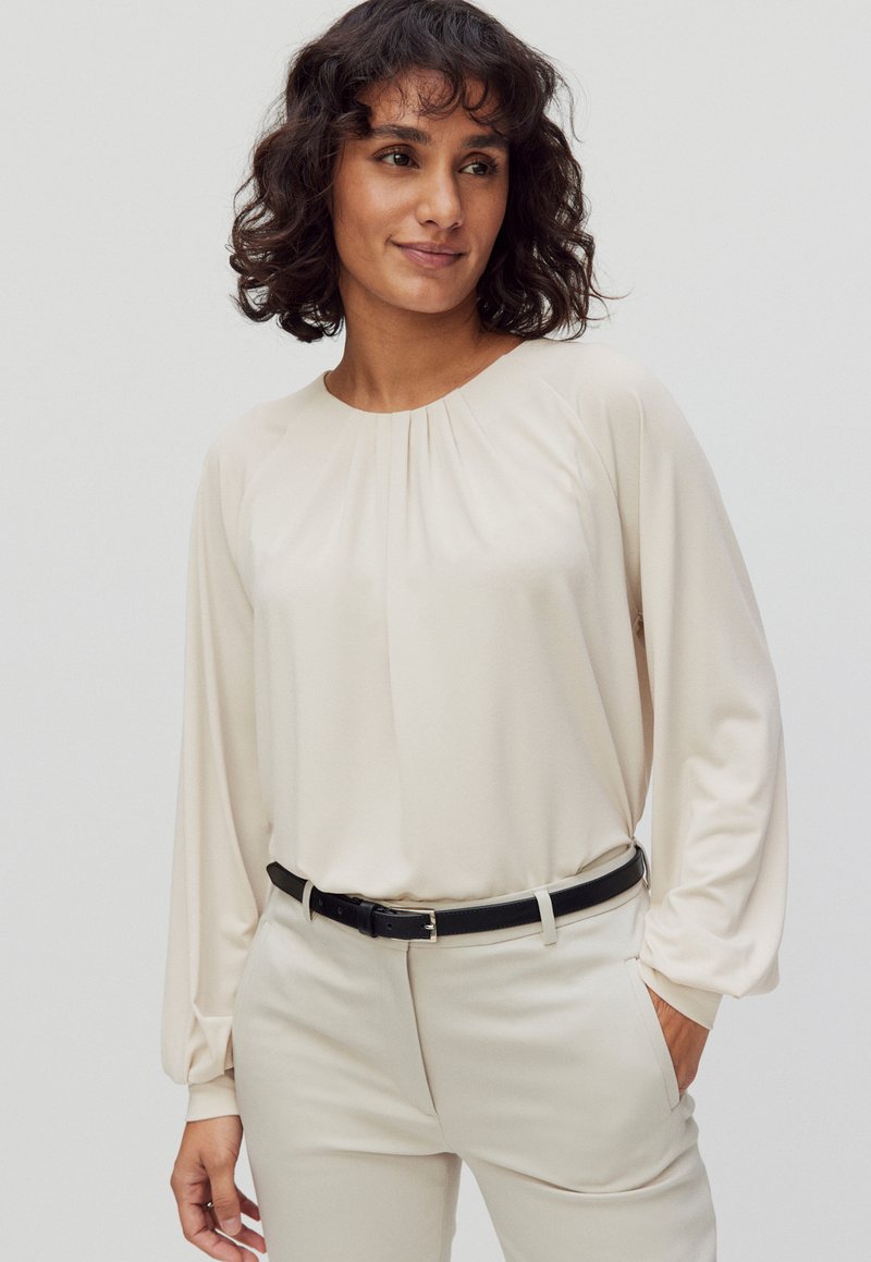 Lyse beige bluse med lange ærmer og samlet detalje ved halsen, parret med beige bukser og et sort bælte. Glat, flydende stof.