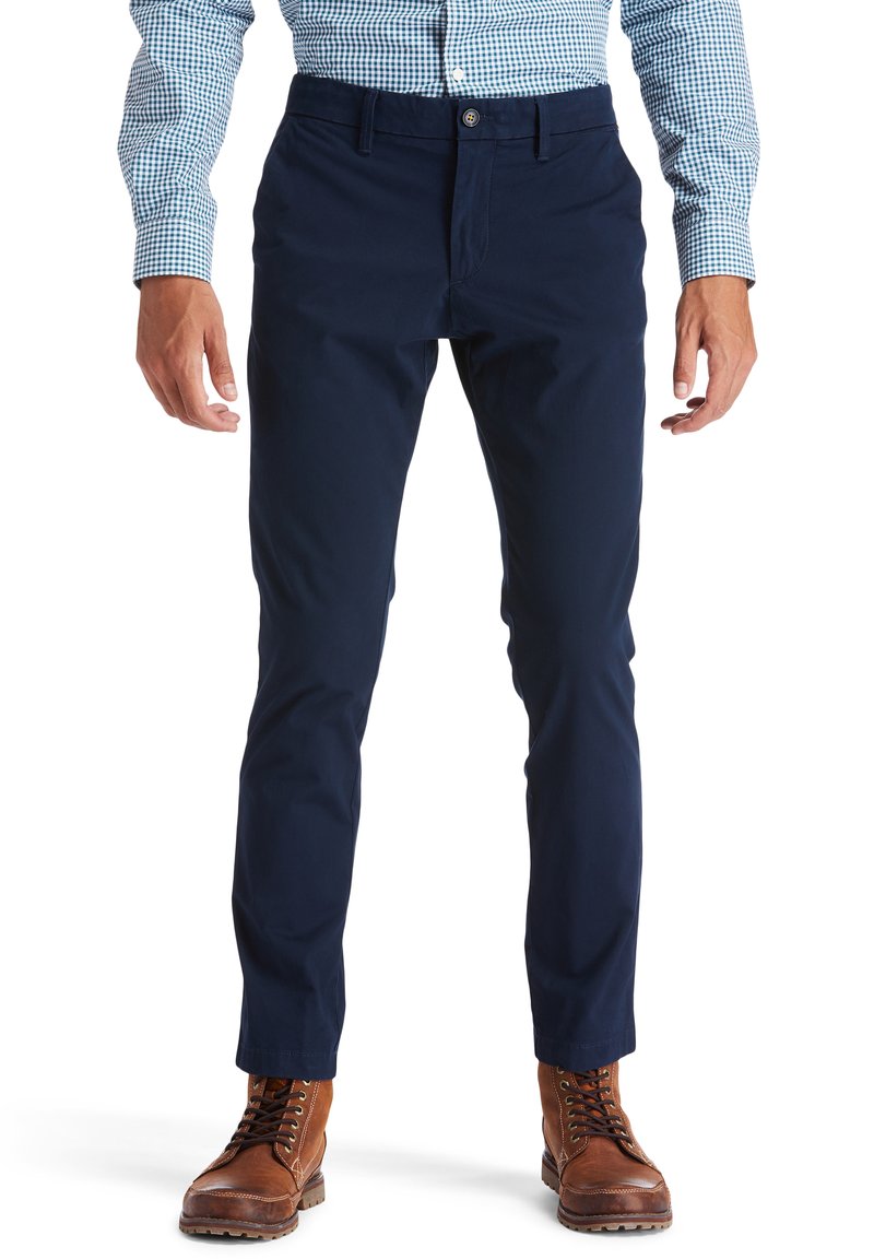 Chinos azul marino hechos de una mezcla de algodón, diseño ajustado, con bolsillos laterales y cierre de botón. Usados con botas marrones.