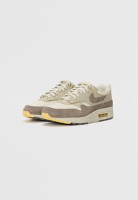 Μπεζ και καφέ αθλητικά παπούτσια Nike Air Max 1 με υλικά από δίχτυ και σουέτ, στρογγυλή μύτη, παχιά καουτσούκ σόλα και ορατή λεπτομέρεια αερόσχης.