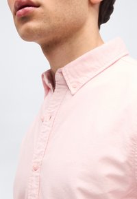 Gros plan sur une personne portant une chemise à boutons rose pâle avec un col, montrant le bas du visage, le cou et le haut de la poitrine.