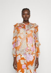 Blusa arancione traslucida con motivi floreali paisley in rosa e bianco, presenta spalline arruffate e chiusura a bottoni sul davanti.