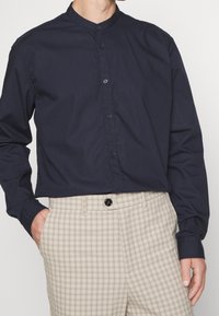 Muž v modré košili s dlouhým rukávem a mandarínským límečkem, se světlehnědými kostkovanými kalhotami s vysokým pasem, s jednou rukou v kapse.