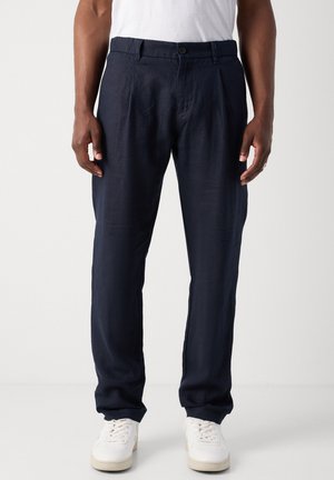 Man som bär marinblå avslappnade byxor och vita sneakers, stående mot en ljus, enfärgad bakgrund.