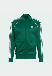 Chaqueta verde con cremallera y cuello alto, hecha de tejido suave. Presenta franjas blancas que bajan por las mangas y un logo bordado en el pecho.