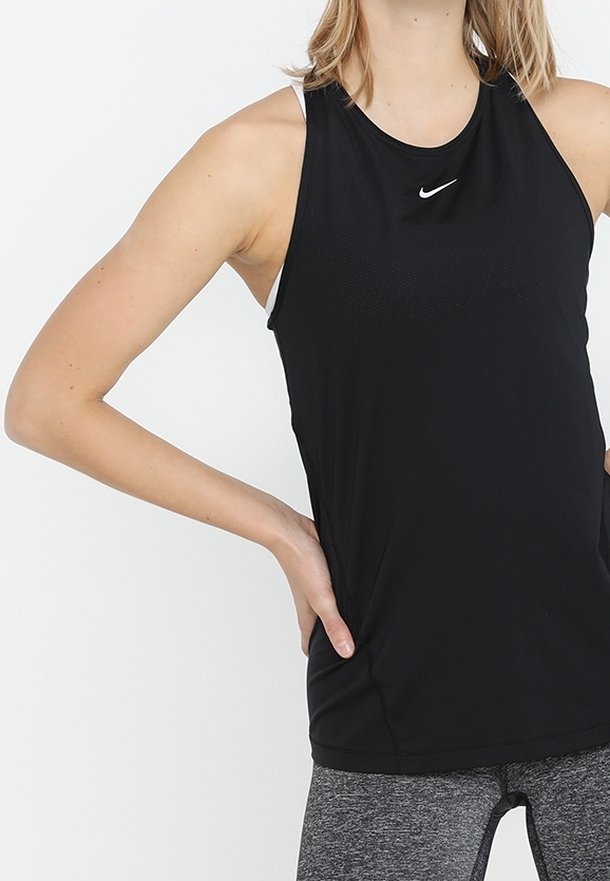 Personne portant un débardeur de sport noir Nike et un legging gris, debout avec une main sur la hanche sur un fond uni.