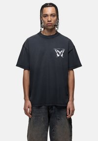 Zwarte oversized katoenen t-shirt met een witte vlinderafbeelding op de linkerborst. De stof heeft een zachte, gladde textuur.