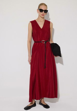 Nő vörös ujjatlan maxi ruhában, fekete övvel, fekete lapos cipőben, napszemüvegben, fehér háttér előtt fekete táskával a kezében.