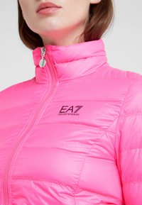 Ljust rosa puffjacka med ståkrage, med kontrasterande svart logotyp och horisontell quiltad textur, metallisk dragkedjedetalj.