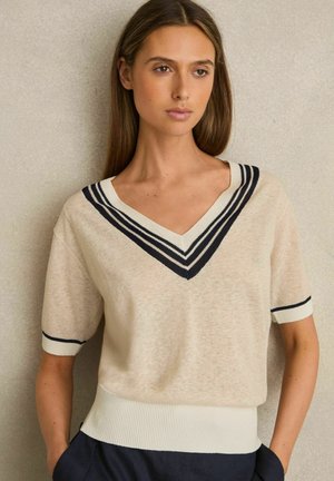 Beige Strickpullover mit V-Ausschnitt. Mit marineblauen vertikalen Streifen um den Kragen und die Bündchen, sowie einem gerippten Abschluss am unteren Rand. Kurze Ärmel.
