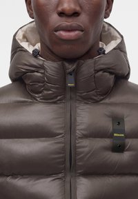 Gros plan d'une personne portant une veste matelassée marron avec capuche, logo Blauer noir et jaune visible sur la fermeture éclair et le patch poitrine.