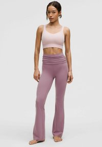 Jasnoróżowy stanik sportowy z cienkimi ramiączkami, sparowany z wysokim stanem, rozszerzającymi się fioletowymi legginsami. Miękka, elastyczna tkanina o gładkiej teksturze.