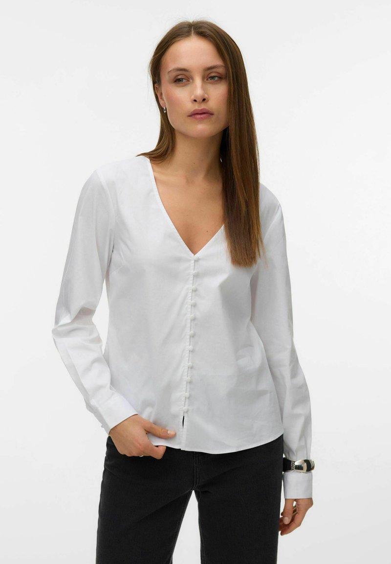 Femme portant un chemisier blanc à manches longues avec col en V et boutons recouverts de tissu, ainsi qu'un pantalon noir, sur un fond uni.