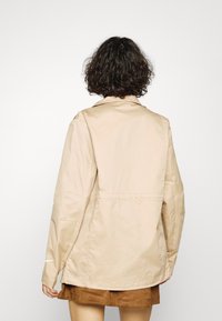 Beige jacket made of smooth fabric, featuring a relaxed fit, long sleeves, and back cinching detail. Minimalist design with no visible patterns.