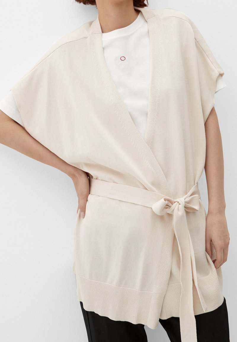 Beige knitted kimono-style cardigan with wide sleeves, a tied belt, and ribbed hem. Worn over a white t-shirt with a minimalist design.