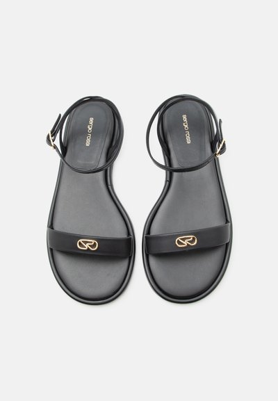 Μαύρα επίπεδα σανδάλια με λουράκια στον αστράγαλο και χρυσή λεπτομέρεια λογότυπου σε φαρδιά μπροστινά λουράκια, τοποθετημένα δίπλα δίπλα σε άσπρο φόντο.