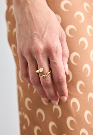 Guld ringe på en hånd, en med cylindrisk form og den anden en glat ring. Brun baggrund med et cremet halvmåne mønster.