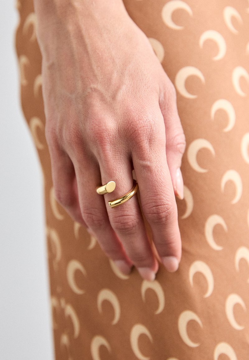 Kuld sõrmused käel, üks silindrilise kujuga ja teine sujuv bänd. Pruun taust sisaldab kreemikat poolkuu mustrit.