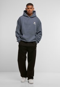 Grå hættebluse med lomme foran og logo, parret med sorte bukser med et unikt accentmønster og hvide sneakers.