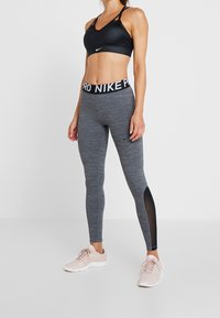 Soutien-gorge de sport noir avec bretelles réglables, associé à des leggings gris avec des panneaux en maille et une ceinture noire. Chaussures de sport rose clair.