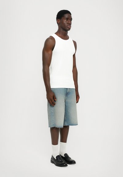 Débardeur blanc sans manches, short en denim bleu clair avec un ourlet effiloché, mocassins noirs et chaussettes blanches, se tenant devant un fond uni.