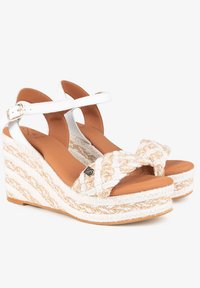 Sandalias de cuña blancas con una parte superior de fibra natural trenzada y una suela texturizada. Cuenta con una correa ajustable en el tobillo y un acento metálico decorativo.