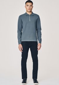 Pelēks zip-up džemperis ar garām piedurknēm, kombinēts ar melnām biksēm un melniem sporta apaviem. Audums ir gluds ar klasisku polo dizainu.