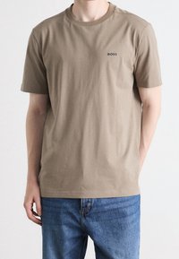 Béžové bavlněné tričko s kulatým výstřihem a krátkým rukávem, na kterém je malá tmavá značka na hrudi. Vytvořeno v kombinaci s modrými džíny.