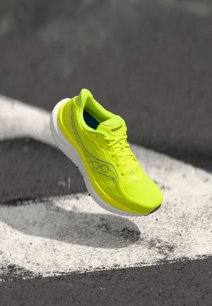 Zapatilla de correr Saucony de color amarillo brillante con suela blanca, en el aire sobre asfalto con una señalización vial blanca.