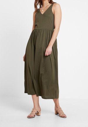 Femme portant une robe midi sans manches vert olive et des sandales à talons blocs beiges ouvertes à l'avant, debout devant un fond blanc uni.