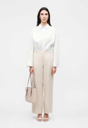 Žena s dlouhými tmavými vlasy nosí bílou pruhovanou košili, béžové kalhoty s širokými nohavicemi, bílé sandály a drží béžovou tašku na dvojitých popruzích.