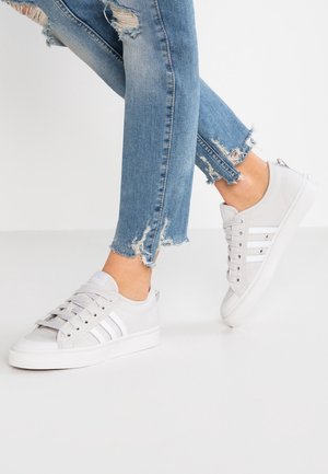 Osoba nosząca jasnoszare trampki z białymi paskami oraz obcięte, niebieskie, podarte rurki, jedna stopa lekko uniesiona.