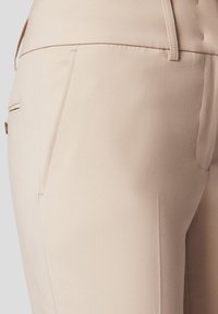 Beżowe spodnie w kroju dopasowanym, wykonane z gładkiego materiału, z bocznymi kieszeniami i subtelnymi detalami w postaci przeszyć wzdłuż szwów.
