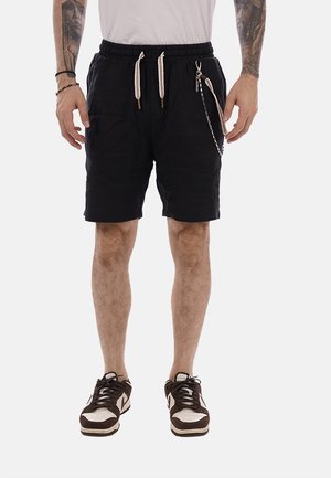 Uomo che indossa pantaloncini neri con coulisse e portachiavi, camicia bianca e scarpe da ginnastica bianconere, mostrando gli avambracci tatuati e i braccialetti.