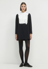 Schwarzes Kleid mit langen Ärmeln, weißem Rüscheneinsatz und Knopfleiste. Kombiniert mit schwarzen Strumpfhosen und glänzenden Schnürschuhen.