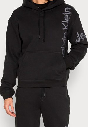 Person in einem schwarzen Kapuzenpullover mit einer Fronttasche, schwarzen Kordeln und vertikalem "Calvin Klein Jeans" Logo auf der rechten Seite.