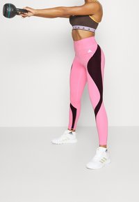 Růžové a černé sportovní legíny s vysokým pasem, vyrobené z hladkého, pružného materiálu a s zakřivenými bočními panely; spolu s hnědou sportovní podprsenkou.