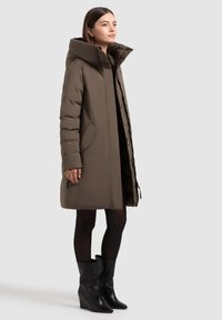 Hnědý zateplený kabát s velkou kapucí, zapínání na zip a bočními kapsami. Modelka v černých kotníkových botách a silonkách. Hladká textura a délka po kolena.