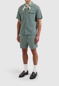 Tweedelig outfit in een gestructureerde groene stof, bestaande uit een kortgevoerd shirt, bijpassende shorts, zwarte instappers en een patroonsjaal.