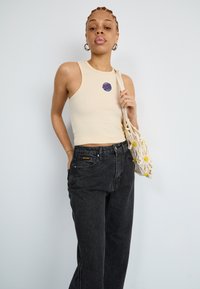 Débardeur côtelé beige clair avec un logo graphique circulaire bleu et rose, associé à un jean noir taille haute et un sac en crochet beige.