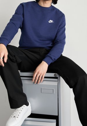 Personne portant un sweat Nike bleu marine, un pantalon noir et des baskets blanches, assise sur un classeur métallique gris sur un fond uni.