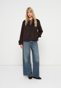 Chaqueta corta marrón con esquinas redondeadas, cuatro botones y puños anchos, combinada con pantalones de mezclilla de pierna ancha en azul claro. Zapatos planos negros.