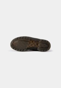 Semelle d’une chaussure marron foncé avec une surface texturée et des sections translucides laissant apparaître une couche intérieure à carreaux, sur un fond blanc uni.