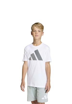 Un garçon porte un tee-shirt blanc Adidas avec un logo gris et un short gris clair, sur un fond blanc uni.