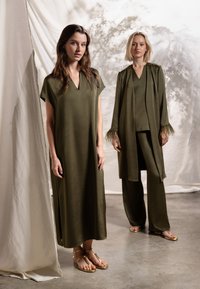 Due donne con abiti verde oliva, una in un vestito senza maniche con sandali, l'altra con un lungo cappotto, top e pantaloni con sandali.