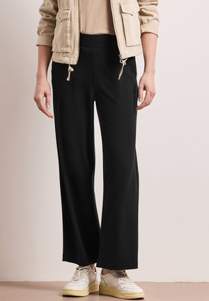 Person trägt beige Jacke, schwarze weite Hose und weiße Sneakers, steht vor einer schlichten, hellen Wand.