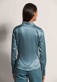 Teal Satin-Hemd mit langen Ärmeln und Kragen, glatte Textur und geknöpften Manschetten. Taillierte Passform mit abgerundetem Saum, kombiniert mit gerippten teal-farbenen Hosen.